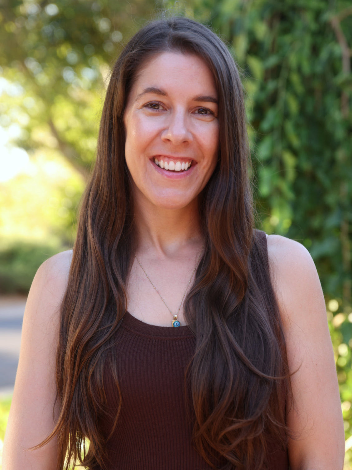 Woman with long brown hair smiling outdoors with greenery in the background