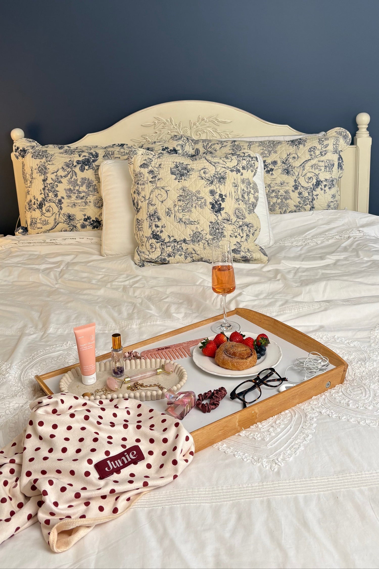 Bed with floral pillows, a tray with a glass of wine and snacks, and a polka dot Junie quick dry hair towel on a dark blue wall background.