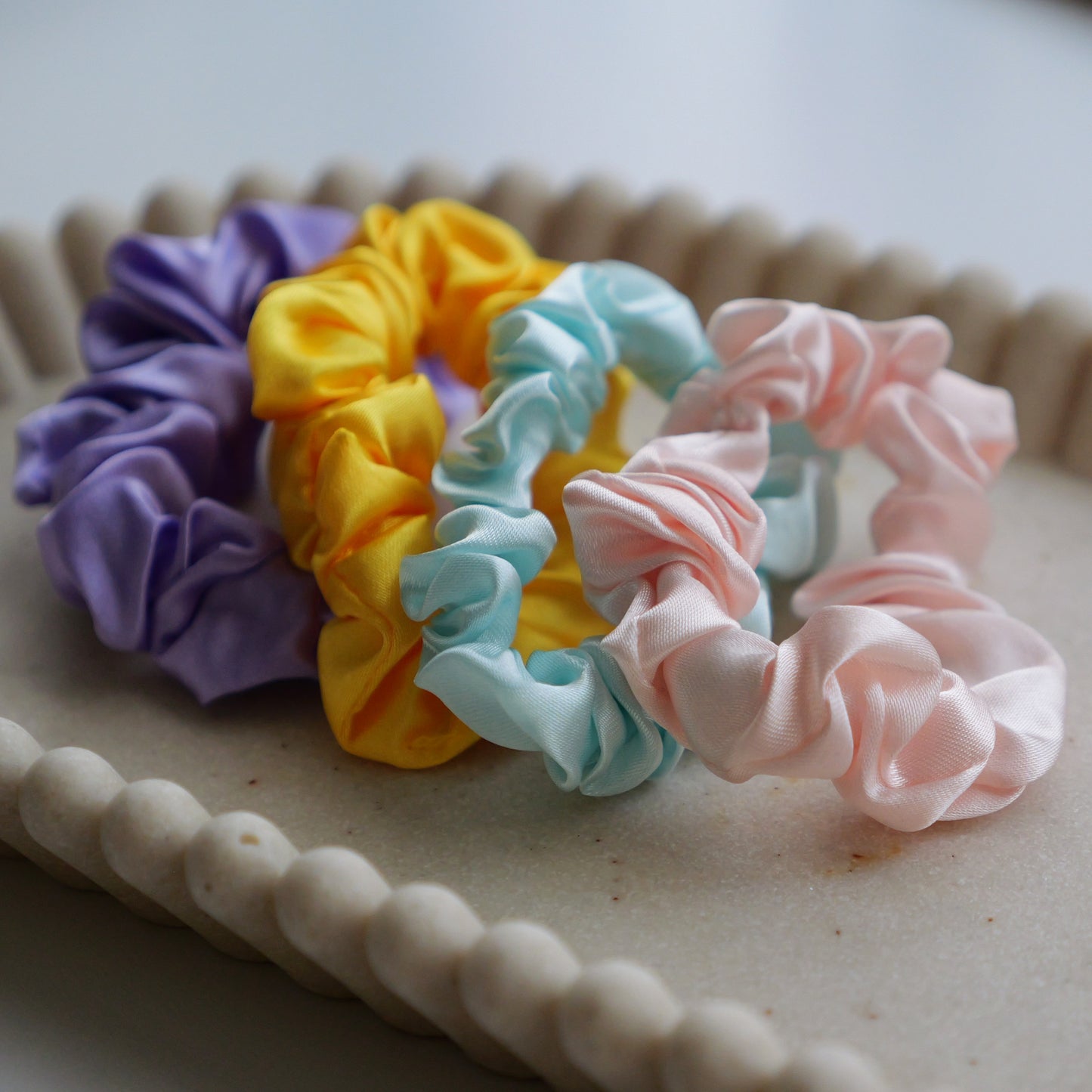 Four silk hair scrunchies in purple, yellow, light blue, and pink, arranged on a beige scalloped tray. kids fairy bread