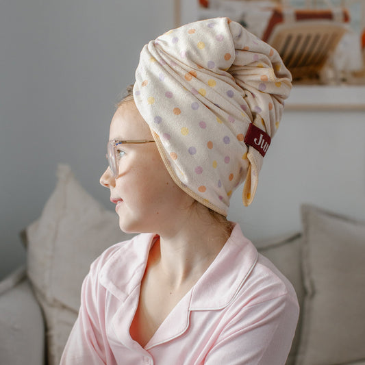 A girl in light pink pyjamas sits on a sofa wearing a Junie quick dry polka dot hair towel with pastel polka dots and a small brown brand tab that reads 'Junie'.