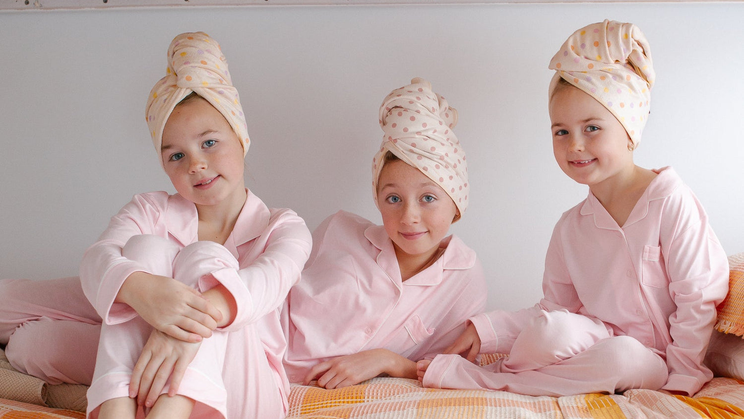 Three children sitting on a bed under a bunk bed, wearing pink pajamas and Junie quick dry hair towel wraps on their heads, with a yellow and white checkered blanket.