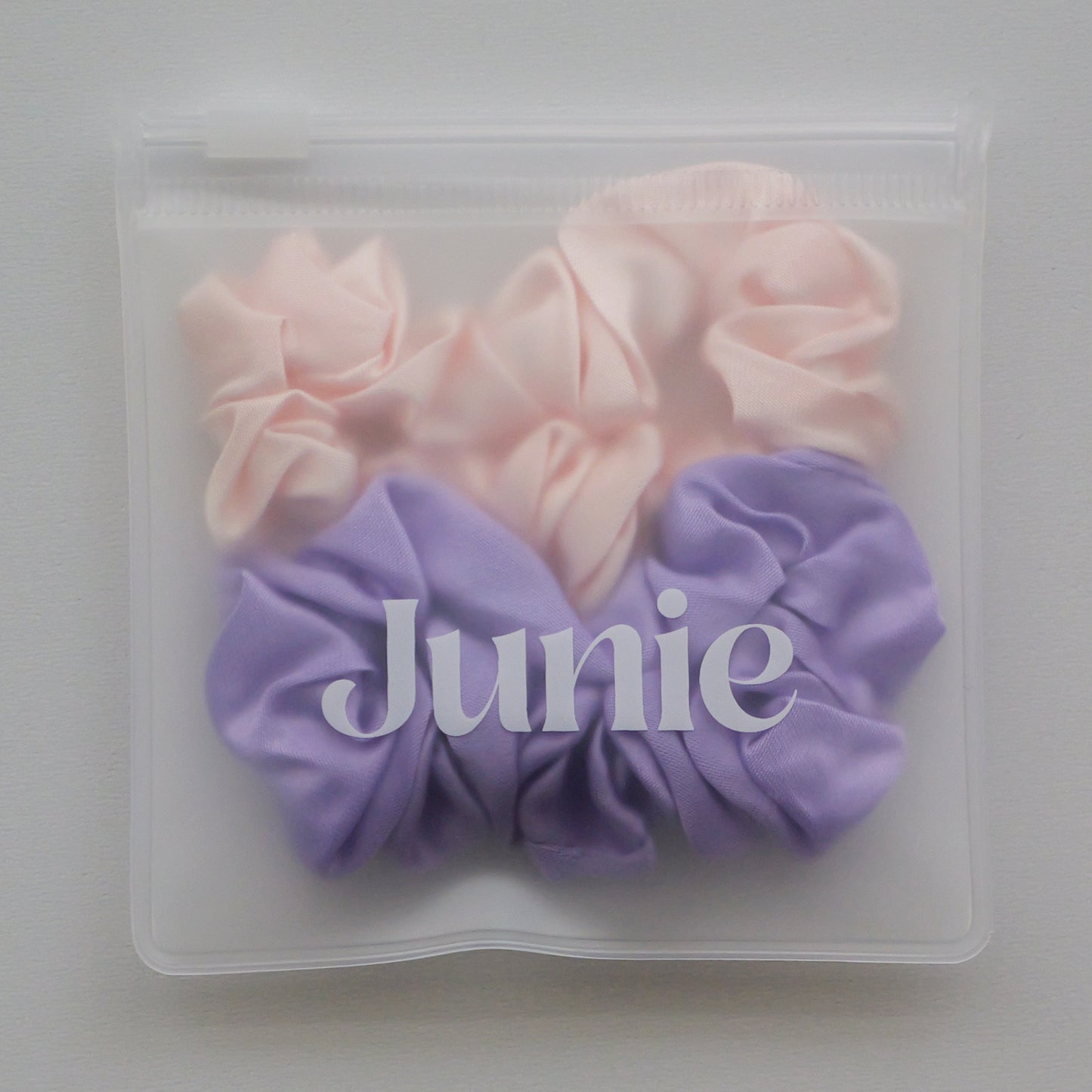 A clear zip-top pouch containing two silk scrunchies, one light pink and one lavender purple, with the word 'Junie' printed across the front.