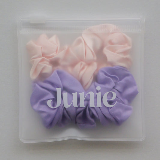 A clear zip-top pouch containing two silk scrunchies, one light pink and one lavender purple, with the word 'Junie' printed across the front.