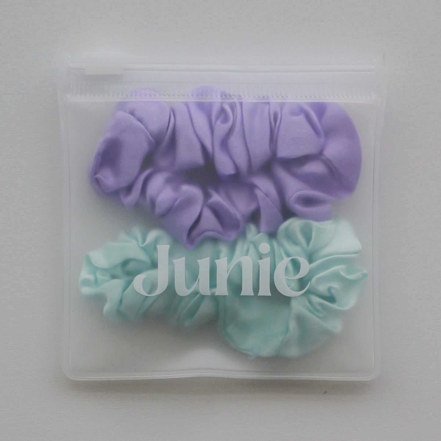 Two silk hair scrunchies, one lavender purple and one baby blue, packaged inside a clear zippered plastic pouch with the brand name 'Junie' printed on the front.