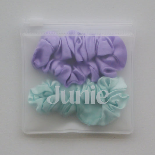 Two silk hair scrunchies, one lavender purple and one baby blue, packaged inside a clear zippered plastic pouch with the brand name 'Junie' printed on the front.