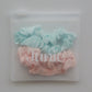 Two silk fabric hair scrunchies, one light aqua blue and one pale pink, packaged inside a clear plastic zip-top pouch with the brand name 'Junie' printed in white across the front.