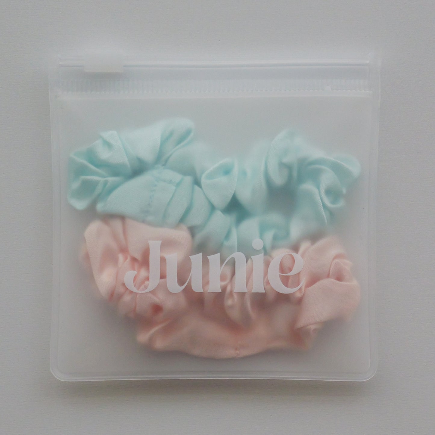 Two silk fabric hair scrunchies, one light aqua blue and one pale pink, packaged inside a clear plastic zip-top pouch with the brand name 'Junie' printed in white across the front.