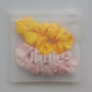 Two silk fabric hair scrunchies, one yellow and one light pink, packaged inside a clear plastic zip-top pouch with the brand name 'Junie' printed on the front.