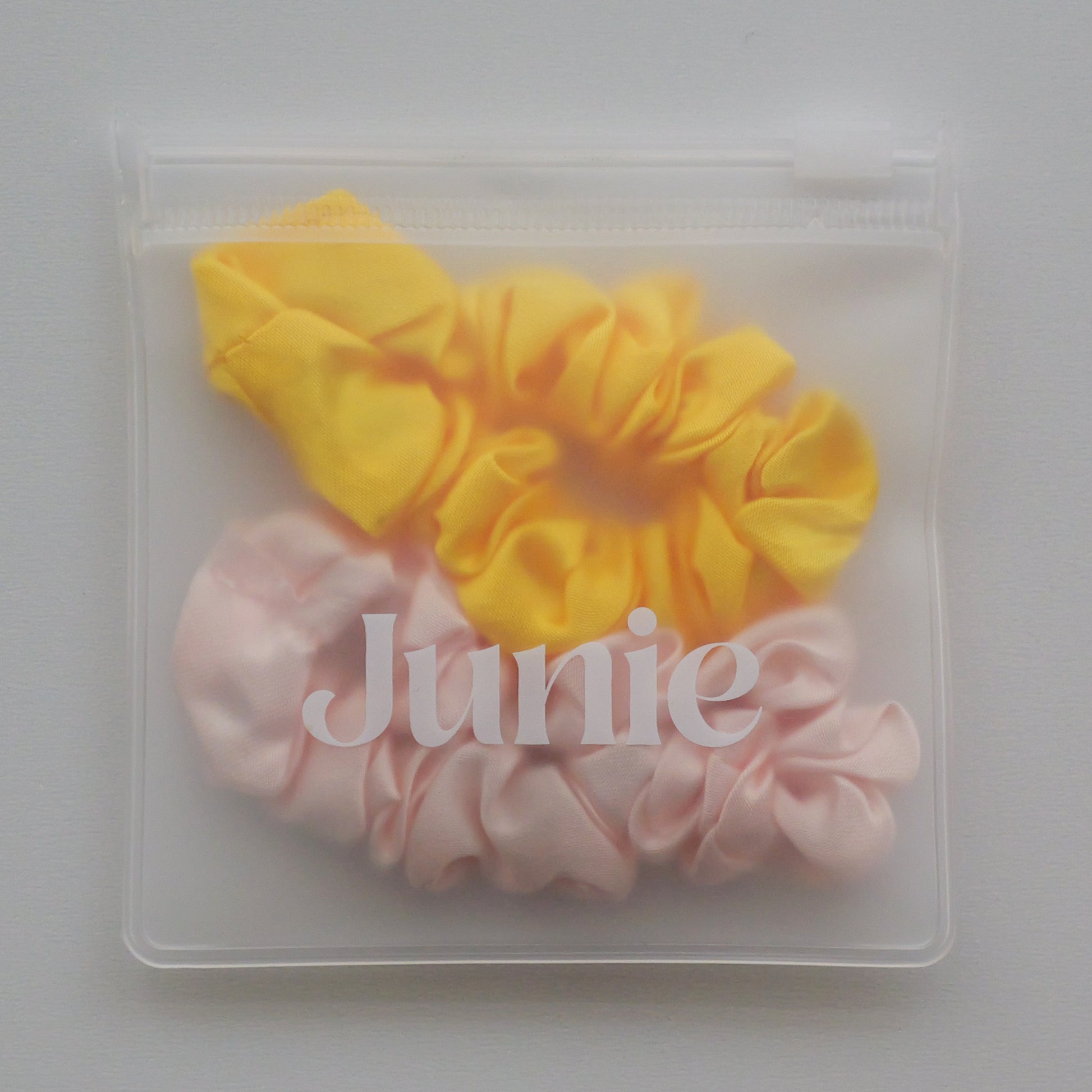 Two silk fabric hair scrunchies, one yellow and one light pink, packaged inside a clear plastic zip-top pouch with the brand name 'Junie' printed on the front.