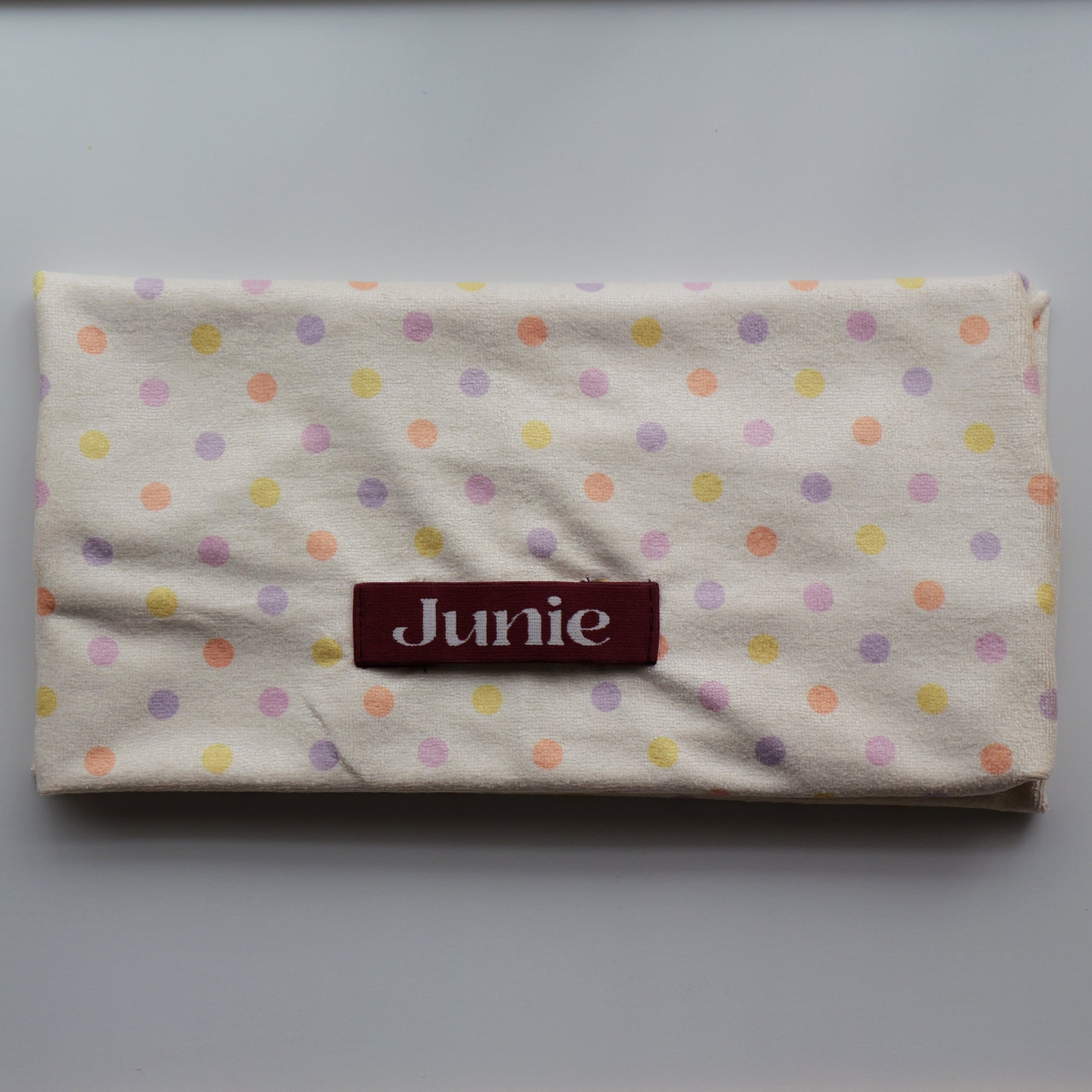 A folded fairy bread cream-colored hair drying towel with a pastel polka dot pattern in pink, purple, yellow, and orange, featuring a dark red rectangular label with the name 'Junie' in white text.