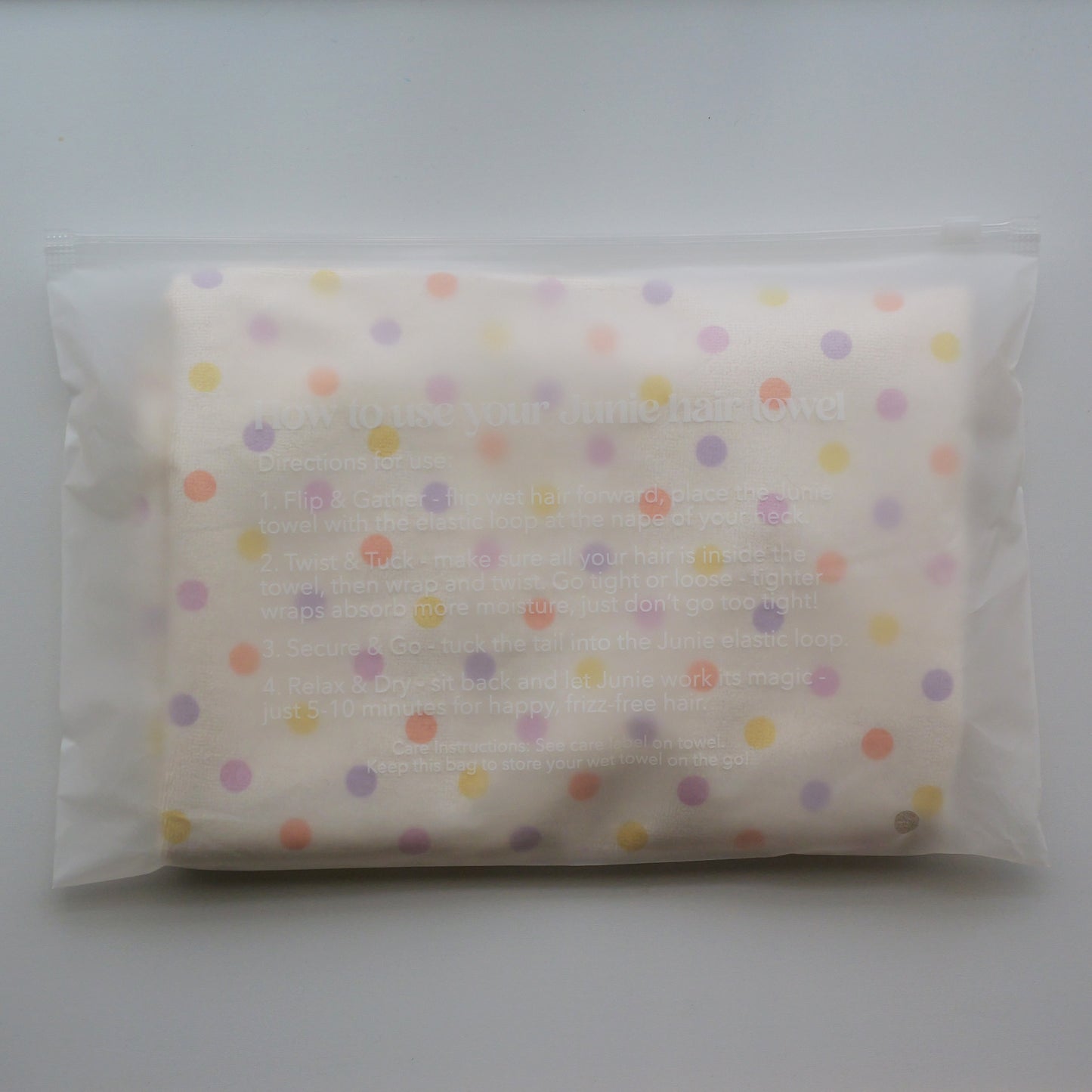 A folded cream-colored Junie quick dry kids hair towel with multicolored polka dots, sealed inside a clear plastic bag with printed instructions on the front.