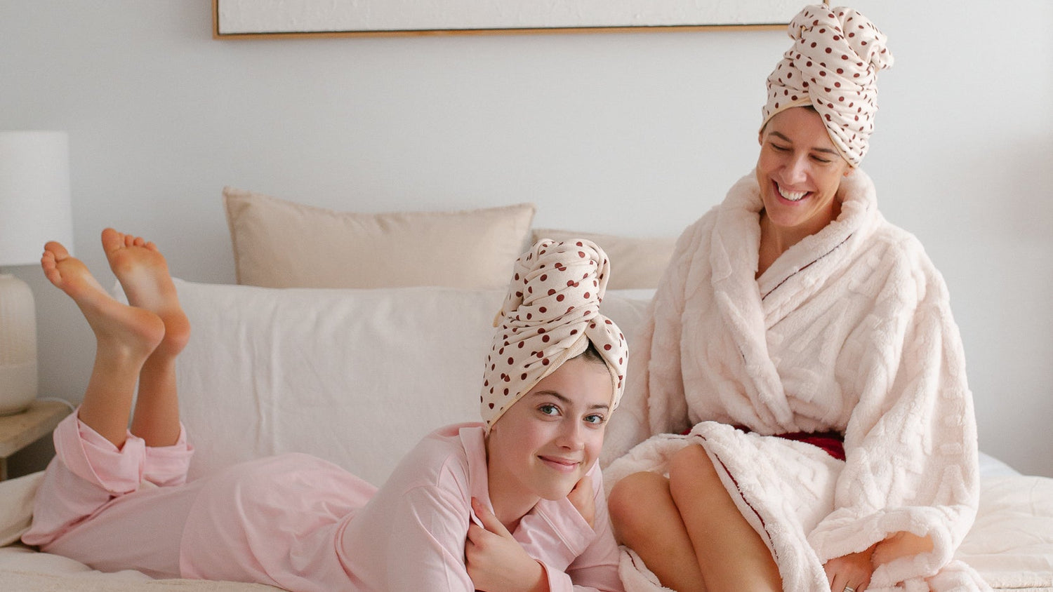 Two women in pink and white robes sitting on a bed with Junie quick dry hair towels on their heads, with a framed art print on the wall behind them.