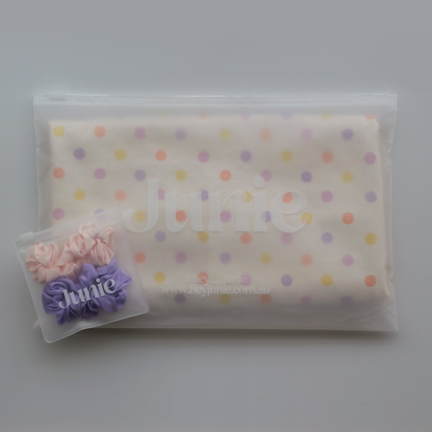 A folded cream-coloured Junie quick dry hair towel kids item with pastel multicolor polka dots, sealed inside a clear plastic zip pouch. A small clear plastic bag containing two silk scrunchies, one pink and one purple, sits in the lower-left corner.