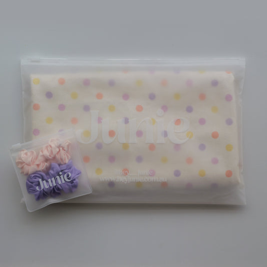 A folded cream-coloured Junie quick dry hair towel kids item with pastel multicolor polka dots, sealed inside a clear plastic zip pouch. A small clear plastic bag containing two silk scrunchies, one pink and one purple, sits in the lower-left corner.
