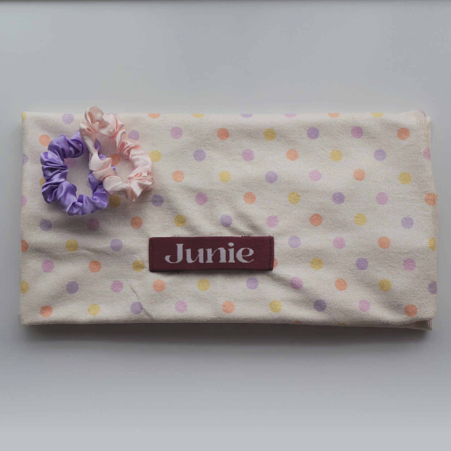 A fabric quick dry hair towel with a cream background and pastel polka dots in pink, purple, and yellow, featuring a maroon label that reads 'Junie' and two silk scrunchies on top.