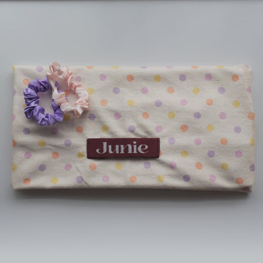A fabric quick dry hair towel with a cream background and pastel polka dots in pink, purple, and yellow, featuring a maroon label that reads 'Junie' and two silk scrunchies on top.