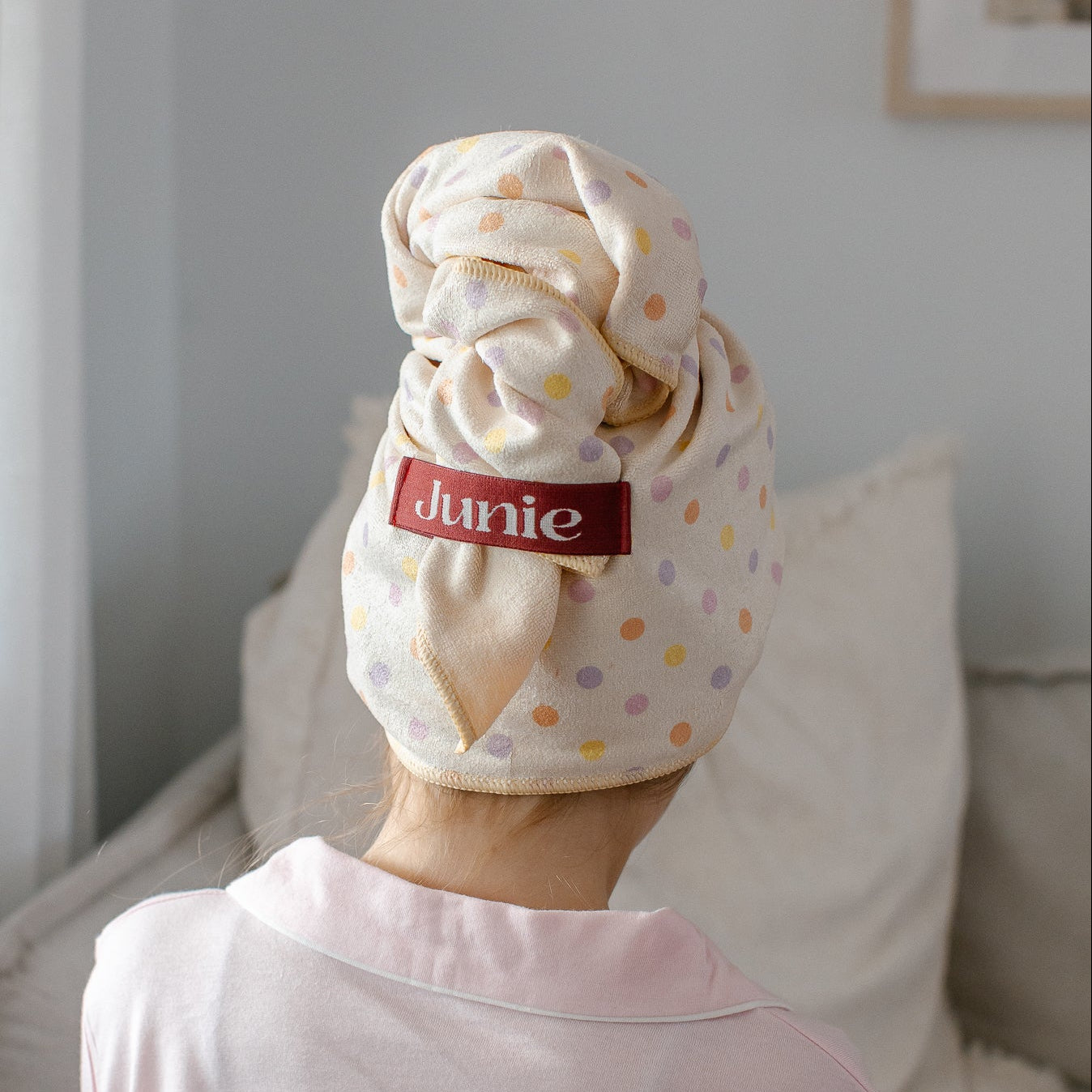 A girl sitting on a sofa indoors, shown from behind wearing a Junie quick dry hair towel with small multicolor polka dots and a red brand label that reads 'junie'.