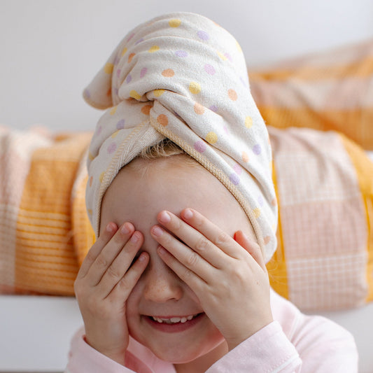 A young child wearing a light pink pyjama shirt and a Junie quick dry hair towel towel with pastel polka dots, covering their eyes in a playful pose in front of a bed with yellow and pink bedding.