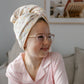 A young girl wearing a light pink pajama shirt and a cream-colored polka-dot Junie quick dry hair towel wrapped around her head, sitting on a gray sofa in a brightly lit room.