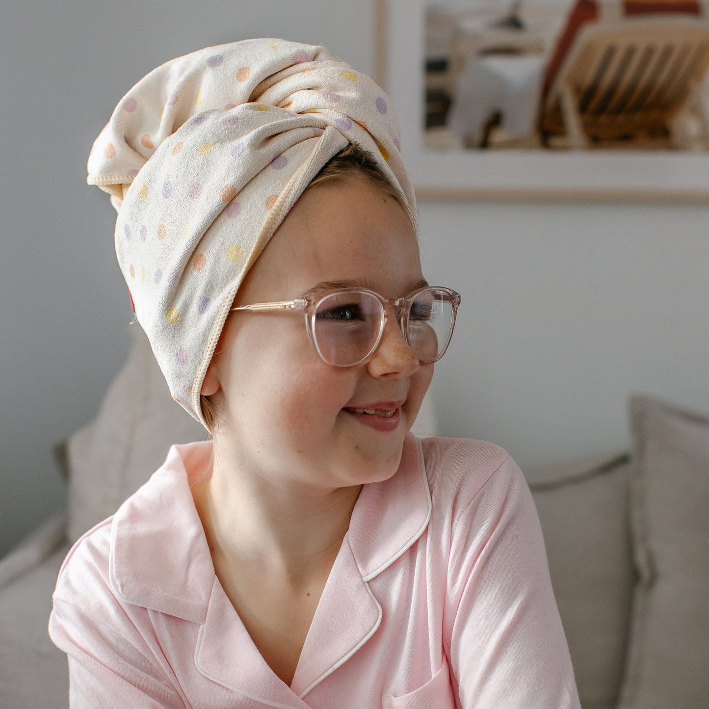 A young girl wearing a light pink pajama shirt and a cream-colored polka-dot Junie quick dry hair towel wrapped around her head, sitting on a gray sofa in a brightly lit room.