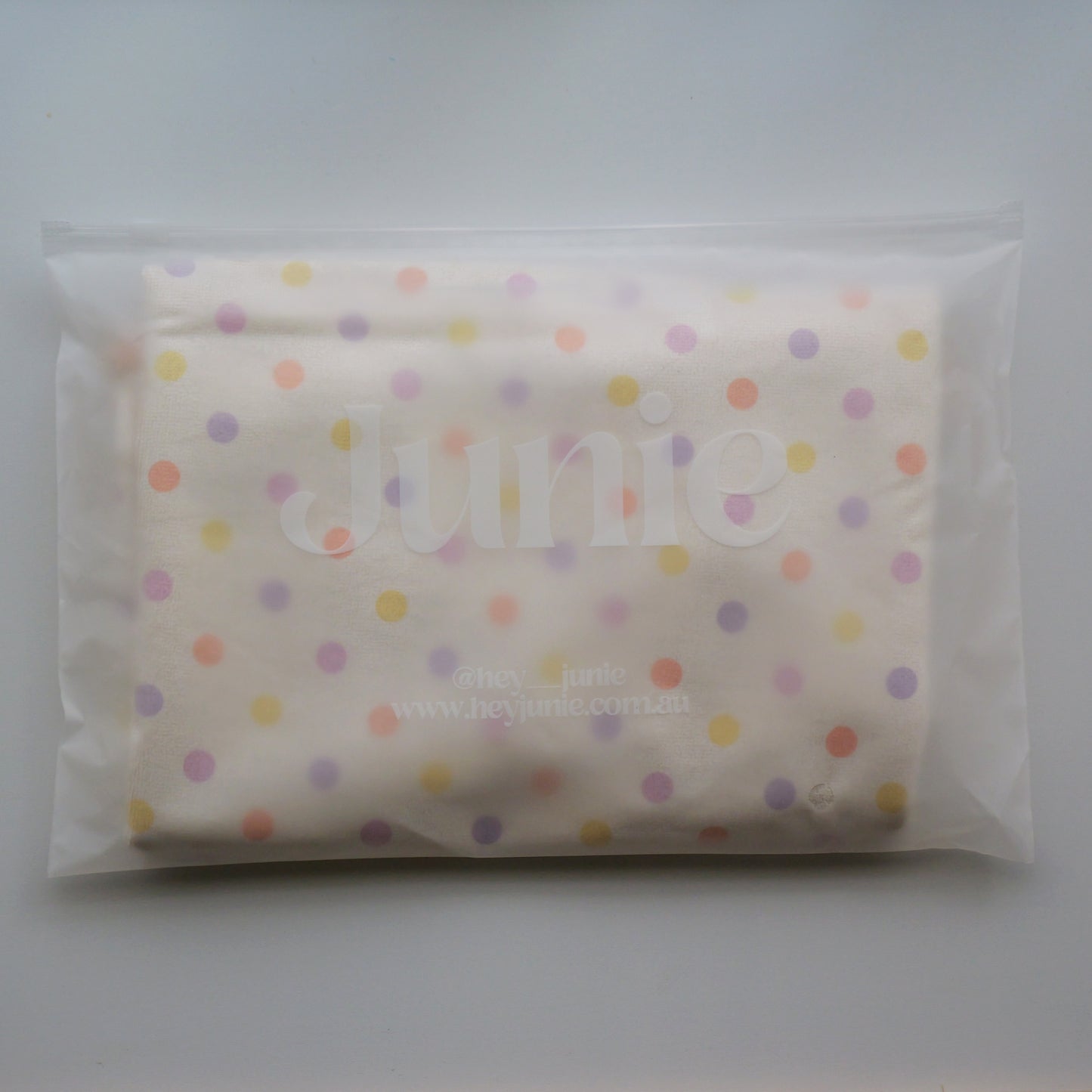 A folded white Junie quick dry kids hair towel, decorated with a pastel polka dot pattern in yellow, orange, purple, and pink. The item is sealed inside a clear plastic bag with a visible 'junie' brand watermark.