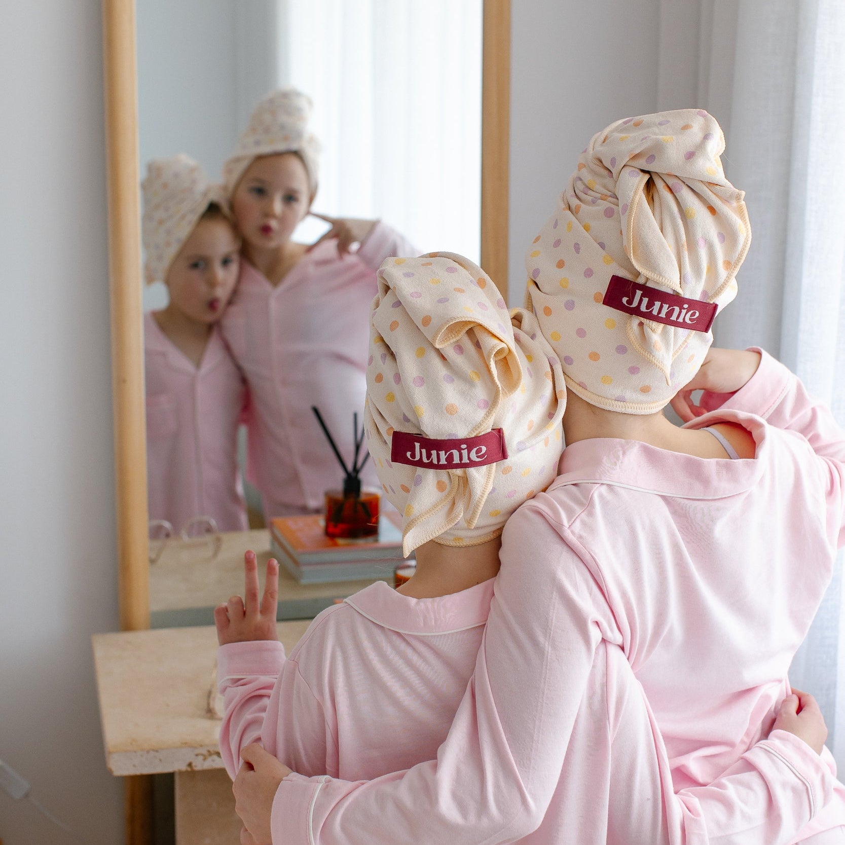 Two girls in matching light pink pyjamas stand in front of a mirror, wrapped in Junie quick dry polka dot patterned hair towels on their heads. The word 'Junie' is printed in red on the bands of their towels.