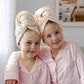 Two young children wearing matching light pink pajamas and having fun with matching Junie quick dry polka dot patterned  pink hair wraps on their heads, sitting together in a brightly lit room.