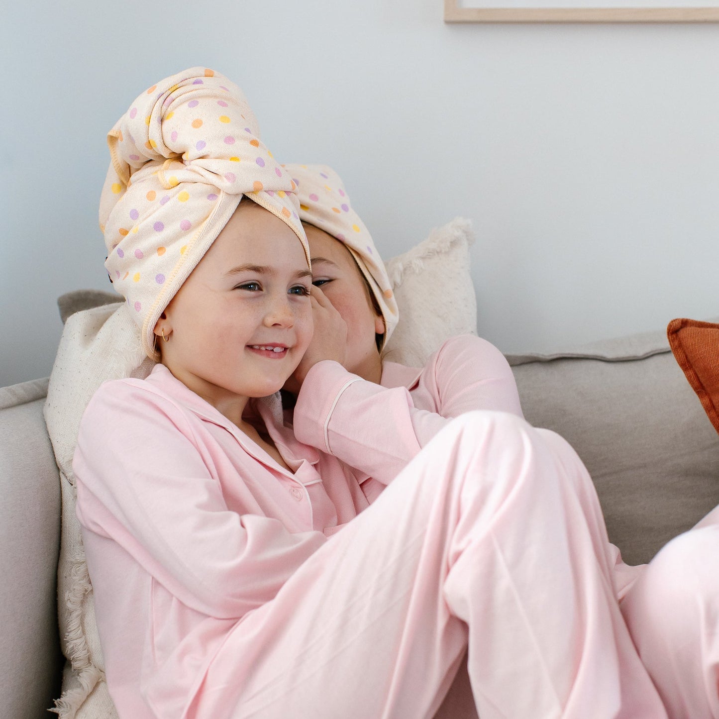 Two children sitting on a light gray sofa wearing pink pajamas and Junie polka dot quick dry hair towels wrapped around their heads, in a brightly lit room.
