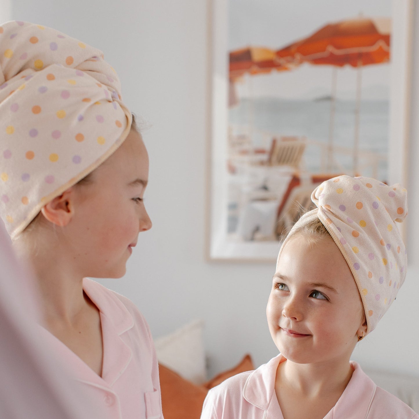 Two children wearing light pink pajamas and matching polka-dot Junie quick dry hair towels wrapped around their heads, standing in a softly lit bedroom.