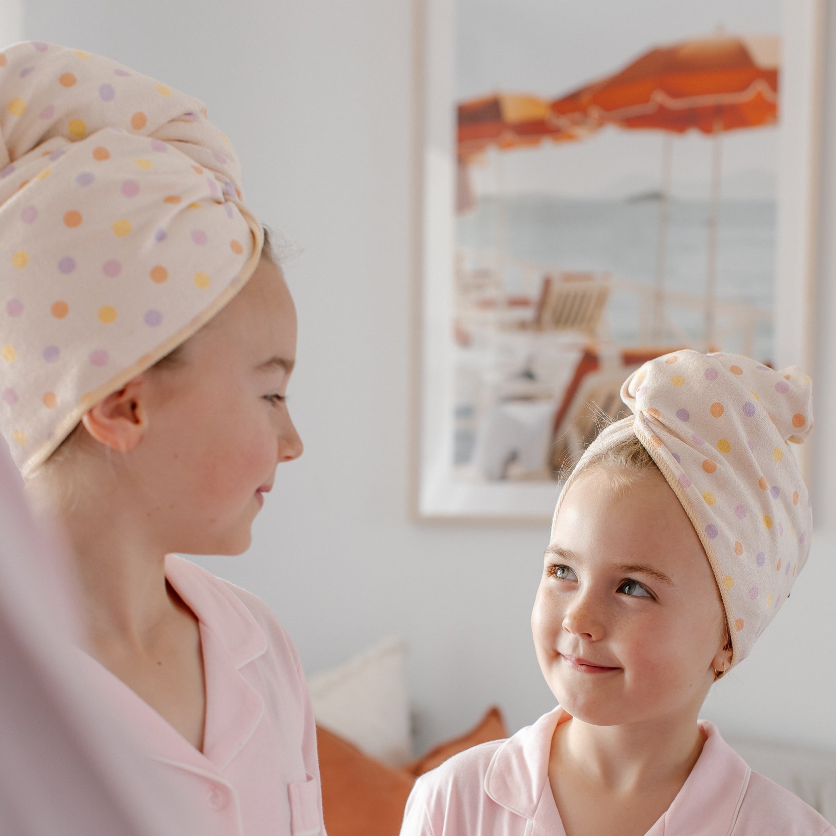Two children wearing light pink pajamas and matching polka-dot Junie quick dry hair towels wrapped around their heads, standing in a softly lit bedroom.